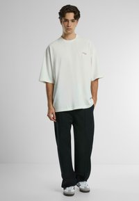 Camiseta de algodón blanca de gran tamaño con cuello redondo y mangas cortas, que presenta un logo impreso sutil en rojo en el pecho. Combinada con pantalones negros.