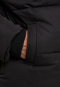 Svart polstret frakk detalj som viser en sidelomme og en svart ribbestrikket mansjett som titter frem fra ermet. Glatt stofftekstur er synlig.