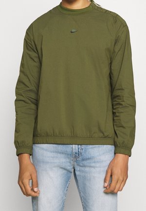 Person iført en olivengrøn langærmet Nike trøje med lille logo på brystet og lyseblå denimjeans, stående med afslappede arme.