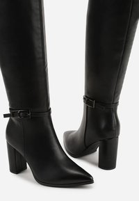 Bottes noires montantes en cuir lisse. Elles présentent un bout pointu, un talon bloc et une sangle boucle réglable près de la cheville.