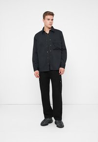 Zwarte button-up shirt met een zak, gedragen over een zwart overhemd met kraag, gecombineerd met zwarte cargobroeken en grijze sneakers. Gladde stof.