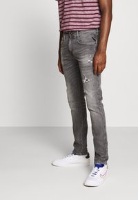 Jeans gris deslavados de corte slim, con detalles desgastados y un diseño clásico de cinco bolsillos, combinados con zapatillas blancas sobre un fondo sencillo.