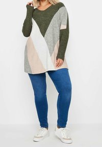 Tunique à manches longues et décolleté en V en vert, gris et rose pâle avec des blocs de couleur géométriques. Portée avec un jean skinny bleu et des baskets blanches.