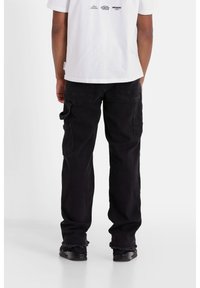 Pantalones cargo negros con múltiples bolsillos, un corte holgado y dobladillos deshilachados, combinados con una camiseta gráfica blanca que presenta varios logotipos.