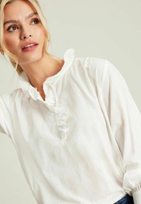 Weißes Blusenshirt mit Rüschensaum, langen Ärmeln und Knopfleiste. Hergestellt aus leichtem Stoff mit einer weichen Textur.