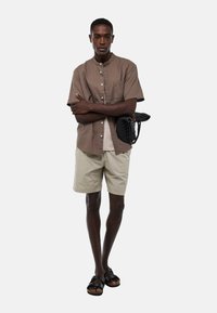 Chemise à manches courtes marron avec un col, shorts beiges et sandales noires. Le modèle tient un sac tressé noir. Fond épuré.