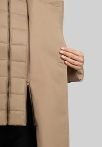 Cappotto imbottito beige con un rivestimento interno trapuntato testurizzato, chiusura con zip e tasche laterali. Tessuto esterno liscio con cuciture a contrasto.
