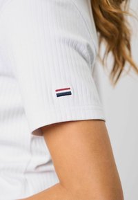 Hvit ribbestrikket t-skjorte med korte ermer, med en liten brodert logo i rødt, hvitt og blått på mansjetten. Glatt tekstur.