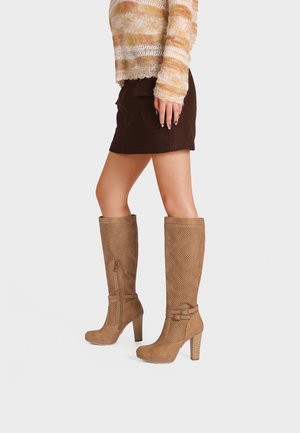 Gambe di una persona che indossa una minigonna marrone, un maglione rigato arancione e beige, e stivali con tacchi alti color cuoio fino al ginocchio con dettagli perforati.