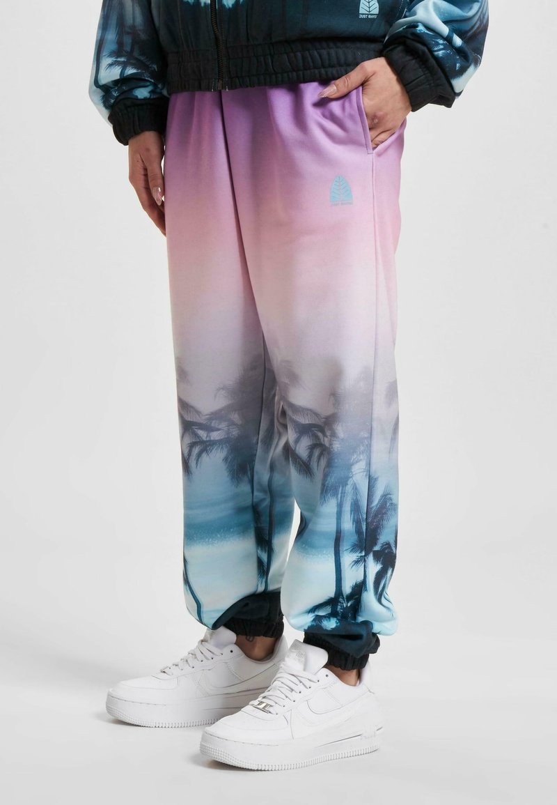 Pantaloni tuta colorati con gradiente rosa e blu, impreziositi da una stampa di palme, dotati di polsini elasticizzati e tasche laterali, abbinati a sneakers bianche.