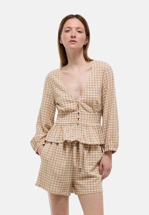 Femme portant un chemisier péplum à manches longues beige et blanc à motif vichy avec boutons et un short taille haute assorti, debout les mains dans les poches.