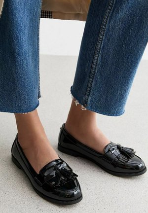 Black patent leather loafers with a round toe, featuring a decorative tassel and subtle stitching details. Worn with frayed denim jeans.