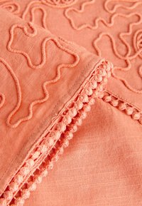 Tissu en corail doux avec des motifs de broderie en relief et une bordure de petits pompons en tissu le long du bord.