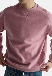 Różowa bluza z teksturowanym wykończeniem, prążkowanym dekoltem w stylu crew oraz subtelnym akcentem z logo. Posiada długie rękawy i luźny krój.