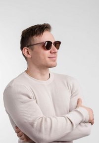 Kulaté sluneční brýle s tmavou skleněnou čočkou a tenkým hnědým rámem. Osoba má na sobě světlý béžový žebrovaný svetr, s překříženýma rukama, mírně otočena na stranu.
