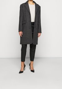 Lång svart och grå rutig kappa med snaggad krage och två framfickor, kombinerad med svarta smala byxor och spetsiga svarta klackar.