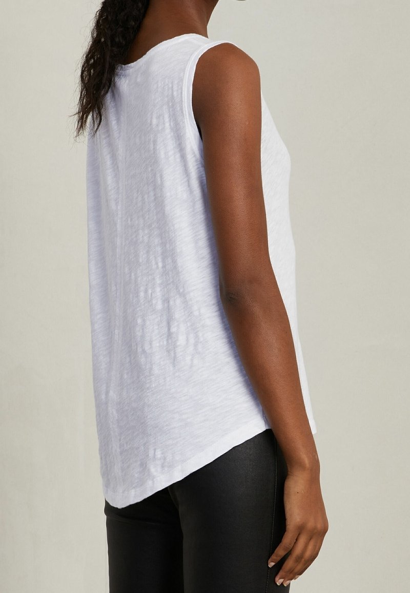 Žena v bezrukávovém bílém texturovaném topu a černých kalhotách, viděná z boku na jednobarevném světlém pozadí.