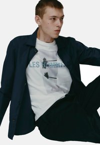 Marineblaues Button-up-Hemd über einem weißen T-Shirt mit Grafik, das Palmen und den Text "LES HOMMES" in Blau zeigt, getragen mit einer dunklen Hose.