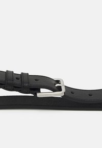 Cinturón de cuero negro con un acabado suave, hebilla de metal plateado, ajustable con varios agujeros, que presenta una sutil inscripción de logo.