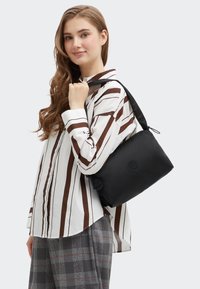 Bolso de hombro de nylon negro con un diseño redondeado, que presenta un logo, correa ajustable y un acabado suave y mate. Camisa a rayas debajo.