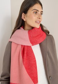 Getextureerde sjaal in roze en rode tinten, met ribpatronen. Het hangt om de nek en complementeert een beige jas en een witte top.
