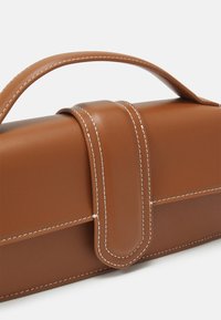 Pochette en cuir marron avec poignée supérieure et rabat avant sécurisé par une lanière cousue, présentée sur un fond blanc.