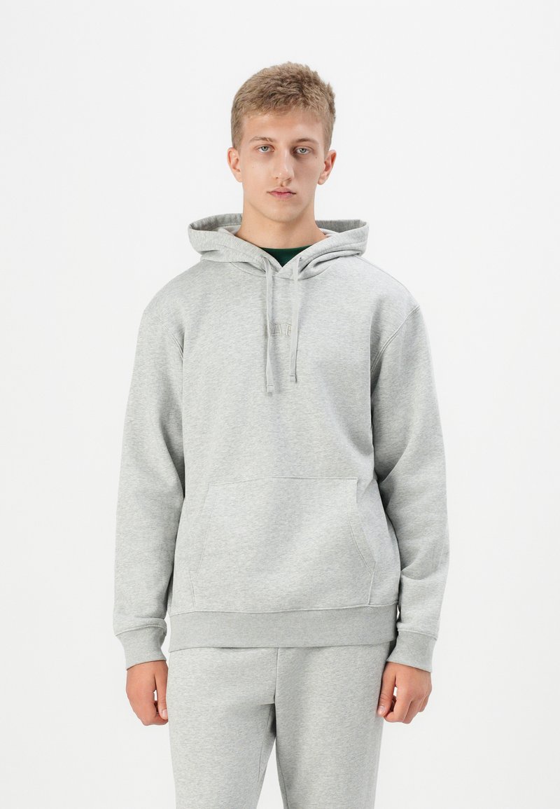 Un sweat à capuche gris en tissu doux avec une poche kangourou, un cordon de serrage et des poignets côtelés. Porté avec un pantalon de jogging gris assorti.