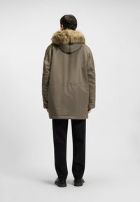 Olijfgroene parka met een nepbont afgewerkte capuchon, zijzakken en een zachte textuur. Draagt over zwarte broek en schoenen.