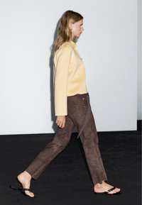 Yellow wool jacket and brown suede pants, with a unique tie detail. The model wears black flip-flops, walking against a neutral background.