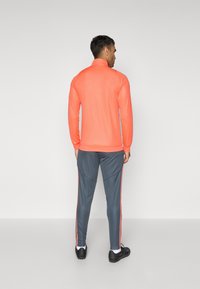 Korallenfarbenes Sportoberteil mit langen Ärmeln und hohem Kragen, kombiniert mit grauen, tapered Hosen mit roten Seitestreifen. Die Schuhe sind schwarz mit weißen Akzenten.