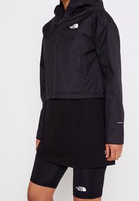 Personne portant une veste noire The North Face avec capuche et un short de cyclisme noir assorti, tous deux arborant les logos de la marque.
