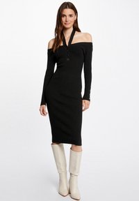 Zwarte geribbelde gebreide jurk met off-the-shoulder ontwerp, knoopsluiting aan de voorkant en lange mouwen, gecombineerd met crèmekleurige kniehoge laarzen.