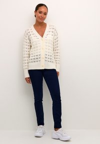 Knitted openwork cream cardigan with a deep V-neck, front buttons, and ribbed cuffs, paired with navy jeans and white sneakers.