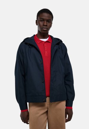 Jeune homme portant une veste à capuche bleu marine par-dessus un pull rouge à col et un pantalon beige, debout devant un fond clair uni.