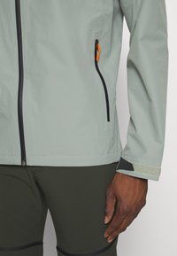 Veste légère grise avec une poche zippée diagonale dotée d'une tirette orange, des finitions noires et des accents réfléchissants au niveau des poignets. Assortie à un pantalon vert.