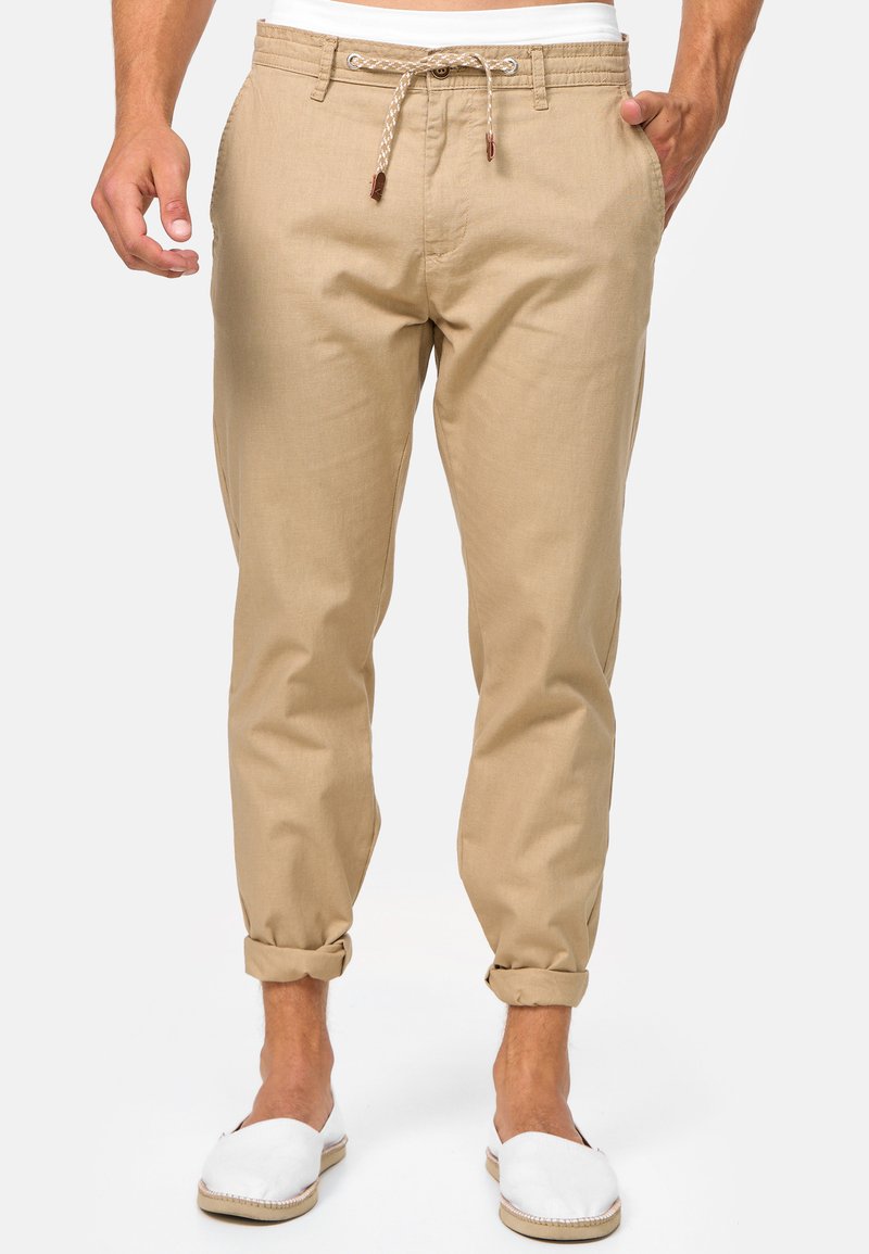 Pantalons en lin beige avec une coupe décontractée, taille avec cordon de serrage et ourlets retroussés. Portés avec des espadrilles blanches. Texture lisse, design minimaliste.
