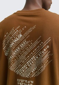 Bruine katoenen t-shirt met witte tekst in verschillende lettertypen en maten, met een ronde ontwerp en geografische coördinaten.