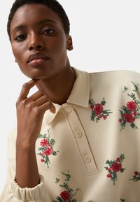 Kremowa polówka z kwiatową haftem w czerwieni i zieleni, z kołnierzykiem i zapięciem na guziki. Materiał o fakturze z marszczonymi mankietami.