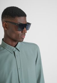 Occhiali da sole neri oversize con design angolare e lenti scure, indossati con una camicia botton-down verde chiaro, caratterizzata da un colletto classico.
