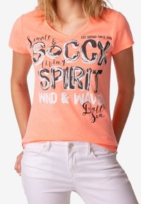 Camiseta de manga corta y escote en V en un brillante color coral con gráficos de texto en blanco y negro, combinada con jeans blancos. Tejido suave, ajuste casual.