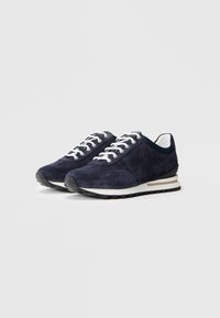 Des baskets en suède bleu marine présentent des lacets blancs, un talon noir contrastant et une semelle intermédiaire blanche, avec une semelle extérieure en caoutchouc texturé pour une meilleure adhérence.