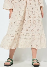 Cremfarb Kleid mit floralen Ausschnitten, gestuften Design und einem Volantsaum. Grüne Sandalen mit gewellten Kanten und einer Schnallendetails.