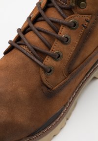 Bota de ante marrón con ojales de metal, cordones marrones y una suela gruesa beige; presenta costuras en contraste a lo largo del borde.