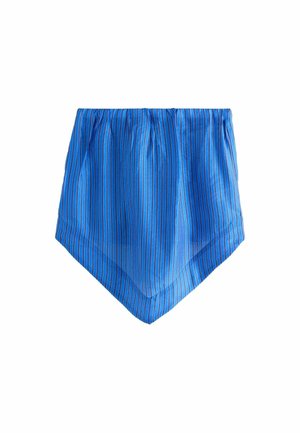 Woman wearing blue strapless jumpsuit and sunglasses standing on rocky beach under pink-striped umbrellas near ocean.