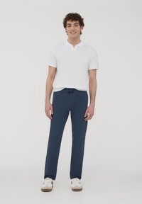 Pantaloni navy con cordoncino realizzati in tessuto morbido, abbinati a una polo bianca a maniche corte. Il modello si trova su uno sfondo neutro.