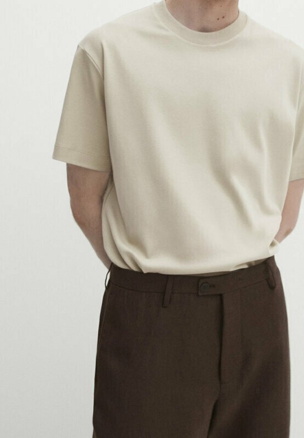 Personne portant un T-shirt beige uni à manches courtes, rentré dans un pantalon taille haute marron foncé avec un bouton et des passants de ceinture.