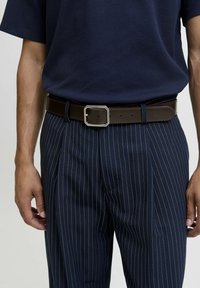 Brauner Ledergürtel mit einer silbernen rechteckigen Schnalle, getragen zu gestreiften marineblauen Hosen und einem strukturierten marineblauen Hemd.
