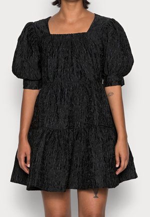 Femme portant une robe courte noire texturée avec des manches bouffantes longueur coude et un décolleté carré, debout devant un fond uni.