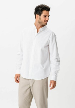 Mężczyzna w białej, długorękawowej, zapinanej na guziki koszuli i beżowych spodniach, patrzący w prawo z delikatnym uśmiechem na tle białego tła.