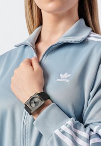 Montre carrée en argent avec un cadran gris et un bracelet en caoutchouc noir, portée sur une main. Veste zippée bleu clair avec le logo et les bandes blanches Adidas.